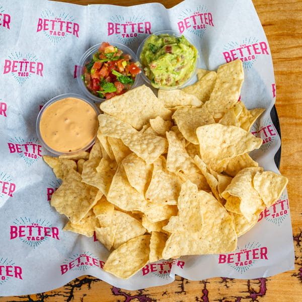 A basket of tortilla chips with sides of salsa, guacamole, and queso from The Better Taco.