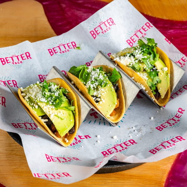 Three The Better Taco tacos with avocado, cilantro, and cheese in a metal holder on branded paper.