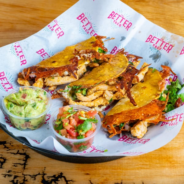 Three crispy chicken tacos with melted cheese, guacamole, and pico de gallo on branded paper.