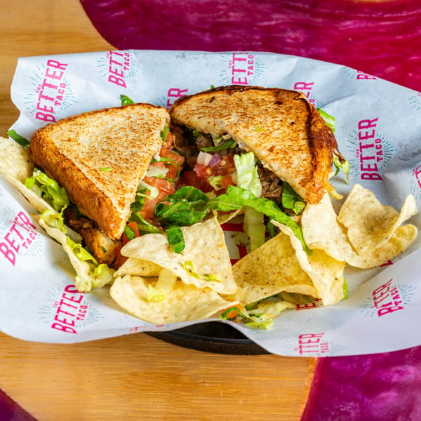 A Better Taco sandwich, cut in half, with lettuce, tomato, and tortilla chips on branded paper.