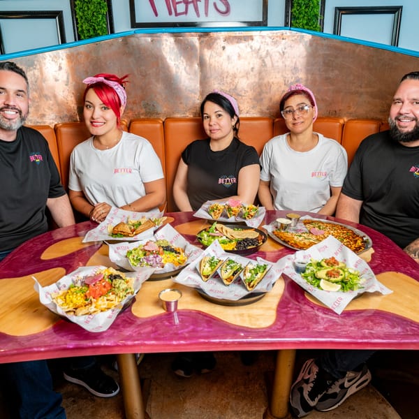 Five smiling staff members of The Better Taco sit around a table laden with various Mexican dishes.