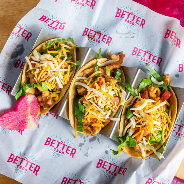 Three "The Better Taco" tacos in a metal holder, filled with meat, lettuce, tomatoes, and cheese.