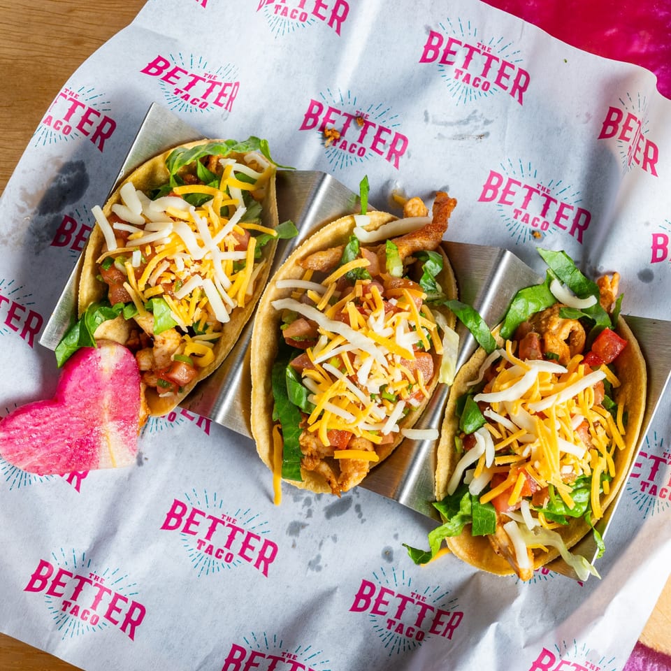 Three "The Better Taco" tacos in a metal holder, filled with meat, lettuce, tomatoes, and cheese.