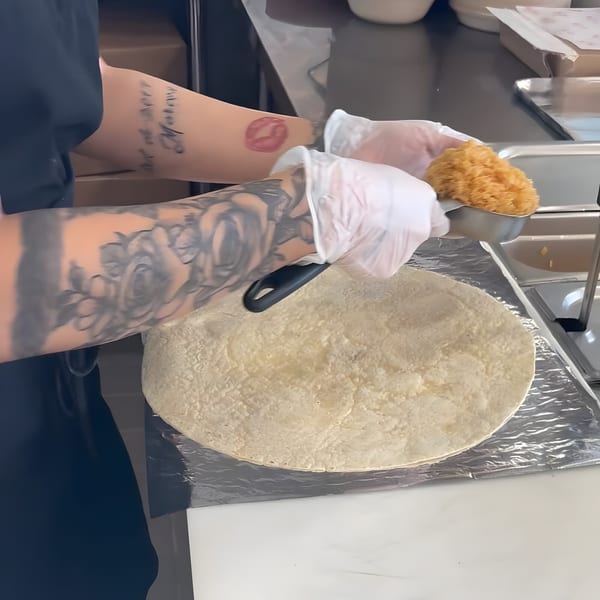 Gloved hands of a person with tattooed arms preparing a burrito on a tortilla.