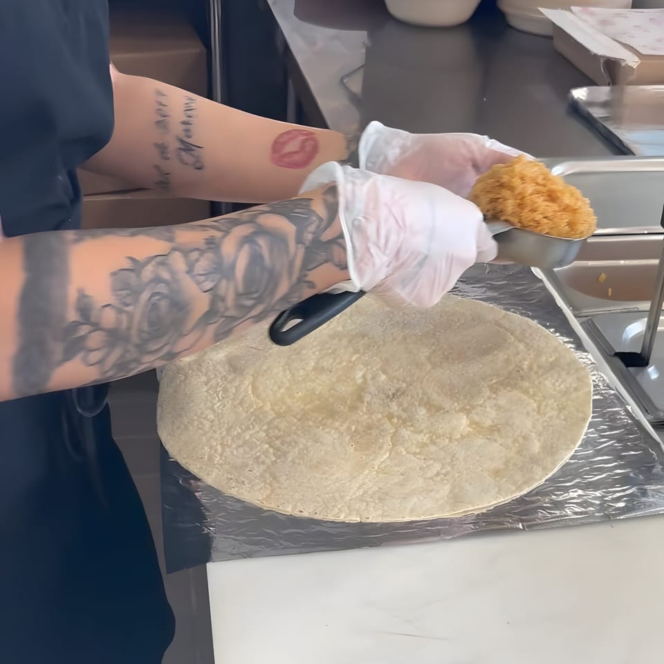 Gloved hands of a person with tattooed arms preparing a burrito on a tortilla.